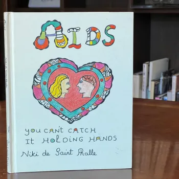 A book titled "AIDS: You Can't Catch It Holding Hands" by Niki de Saint Phalle stands on a wooden table, featuring a colorful illustrated cover with two faces inside a heart.