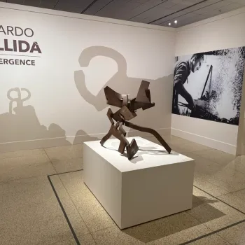 Gallery exhibit of Eduardo Chillida’s sculpture, titled "Convergence," features a metal abstract artwork on a pedestal, wall text, and a black-and-white photo of the artist in the background.