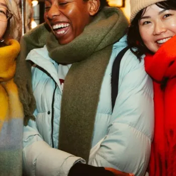 Three people wearing winter coats and colorful scarves stand closely together indoors, smiling and looking at the camera.