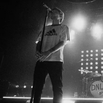 A black-and-white photo shows a singer at a microphone, a keyboardist, and a drummer performing on a stage with bright lights in the background.