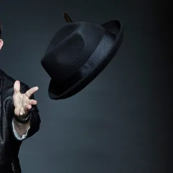 A man in a leather jacket and plaid shirt tosses a black fedora hat in the air against a dark background.