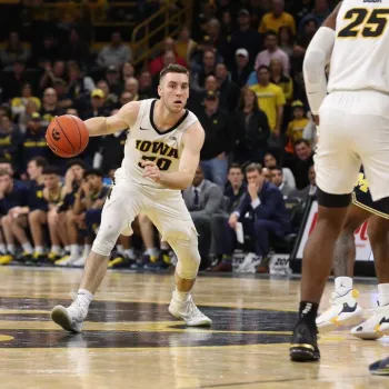 A basketball player in a white Iowa uniform dribbles the ball while being guarded by players in Michigan uniforms on an indoor court.