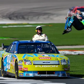 A race car driver does a backflip next to a blue and yellow Ford Fusion stock car on a racetrack, as a crew member stands nearby.