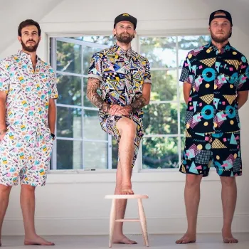 Three men stand barefoot in a bright room wearing colorful, patterned short-sleeve shirts and shorts; one rests his foot on a stool in front of a window.