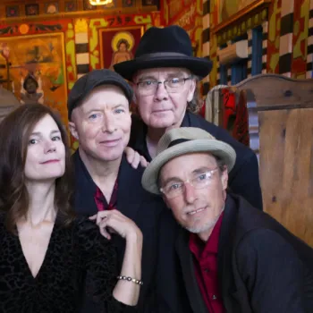 Six adults pose indoors, dressed in dark clothing, standing closely together and looking at the camera with neutral expressions. Colorful decor is visible in the background.