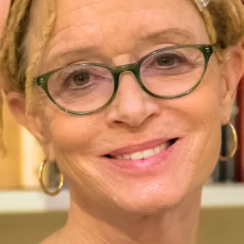 A woman with glasses, hoop earrings, and dreadlocks smiles in front of a bookshelf filled with books.