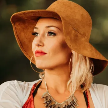 Woman with blonde hair wearing a wide-brimmed brown hat, hoop earrings, and a statement necklace, looking to the side with a neutral background.