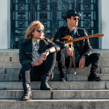 Two men in sunglasses and black outfits sit on outdoor stone steps; one holds an electric guitar. A large building with ornate doors is in the background.