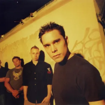 Four men stand in front of a graffiti-marked wall, with one man in the foreground looking directly at the camera under yellow lighting.