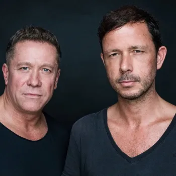 Two men with short hair pose against a dark background, both wearing dark-colored shirts and looking directly at the camera.