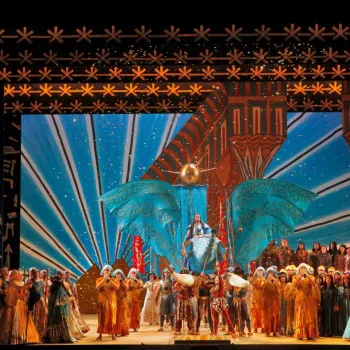 A large group of performers in elaborate costumes stand on a colorful, Egyptian-themed stage with ornate backdrops and props during an opera production.