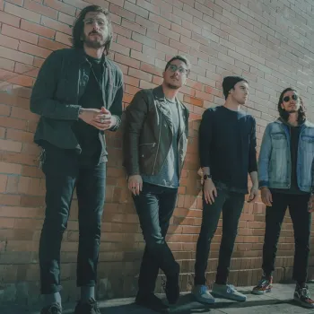 Four men stand against a brick wall, wearing casual jackets and jeans, looking in various directions.