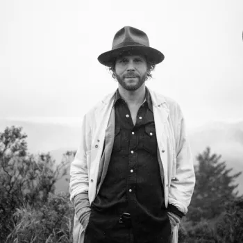 A man in a coat and hat stands outdoors with a foggy, wooded background; another hat appears to be floating or thrown in the air nearby.