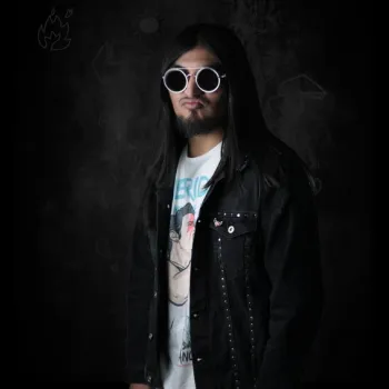 A man with long black hair, wearing round white sunglasses, a black studded jacket, and a graphic T-shirt stands against a dark background.