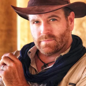 A man wearing a brown wide-brimmed hat, beige shirt, and dark scarf stands indoors by a sunlit stone wall, looking directly at the camera.