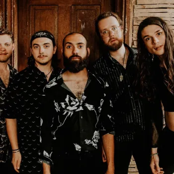 Five people stand in front of a rustic wooden wall, facing the camera. They are dressed in patterned, dark clothing.