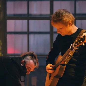 Two people in a warmly lit room; one is tuning or playing an acoustic guitar, while the other person bends over near them. A large window and lamps are in the background.