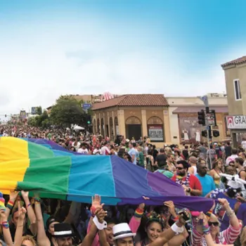 Large crowd participates in a pride parade, holding up a big rainbow flag that stretches over their heads along a city street under a clear blue sky.