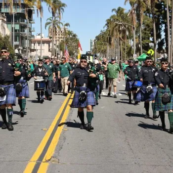 A group of people in kilts play bagpipes and drums while marching down a street in a parade. Spectators and palm trees line the sidewalks.
