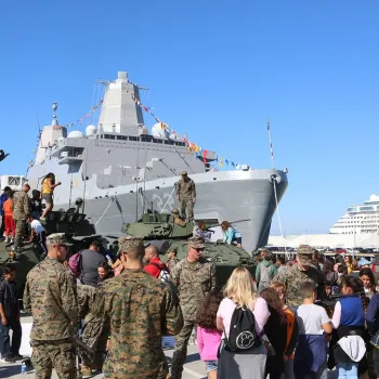 A crowd gathers around military personnel, vehicles, and a navy ship docked at a harbor during a public event.