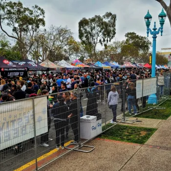 A large crowd gathers behind metal barriers at an outdoor event lined with vendor tents on a cloudy day.