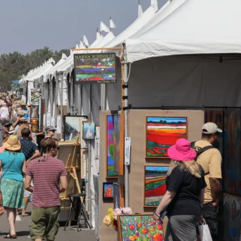 People walk through an outdoor art festival, browsing colorful paintings displayed in white tents along a street lined with palm trees.