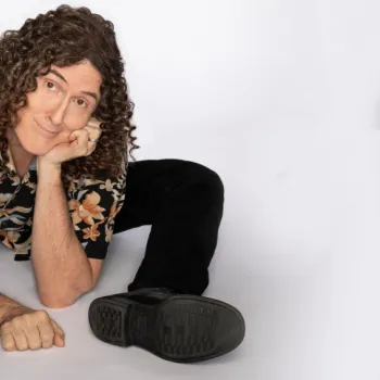 A person with long curly hair in a floral shirt and black pants sits on the floor, legs spread, resting their chin on one hand and looking at the camera.