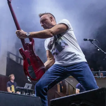 A musician in a white t-shirt and jeans plays a red electric guitar energetically on stage, with smoke and an audience in the background.