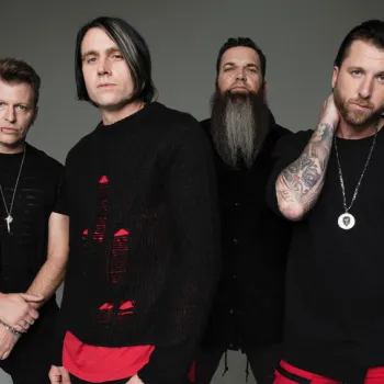 Four men stand together against a plain background, all wearing black clothing with some red accents. One man has long hair and a long beard, while another has tattoos on his arms.