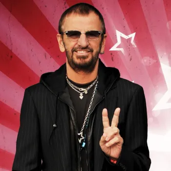 A man in dark sunglasses and layered necklaces stands in front of a red and white starburst background, holding up a peace sign with his right hand.