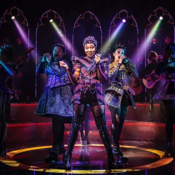 Five performers in elaborate, sparkly costumes sing and dance on stage with colorful lights and musicians in the background.