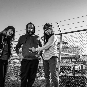 Three people stand together by a chain-link fence; the person in the center wears a hooded jacket, and the person on the right touches their stomach as all three face the camera.
