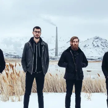 Four men in dark clothing stand in a snowy landscape with mountains, reeds, and a smokestack in the background.