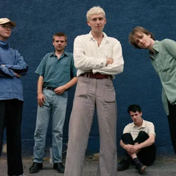 Five young men pose in front of a blue wall, with three standing and one sitting on the ground; all wear casual clothing.
