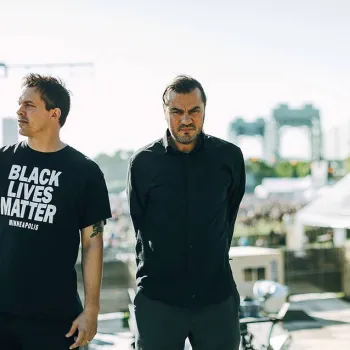 Two men stand on an outdoor stage; one wears a "Black Lives Matter Minneapolis" t-shirt, and the other is dressed in black with hands behind his back. A crowd and bridge are visible in the background.