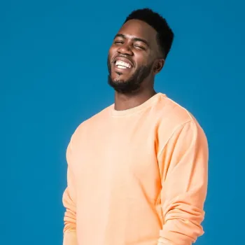 A man wearing a peach-colored sweatshirt stands against a blue background, smiling and looking at the camera.