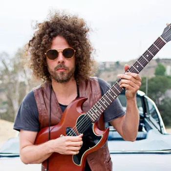 A man with curly hair and sunglasses stands in front of a car, holding a Gibson electric guitar.