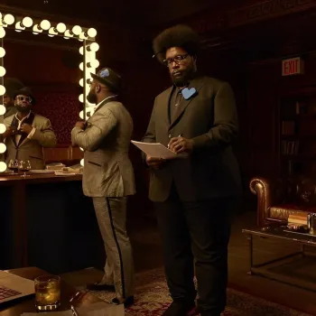 Two men in formal attire are in a dimly lit room; one is standing by a mirror adjusting his hat, while the other writes on a clipboard.