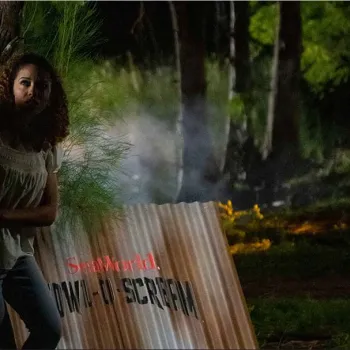 A man and woman stand closely together behind a corrugated metal sign at night in a wooded area, looking cautiously into the distance. Smoke and trees are visible in the background.