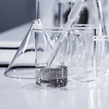A close-up of glass beakers and a conical flask on a laboratory table, with a liquid-filled beaker holding a plastic pipette.