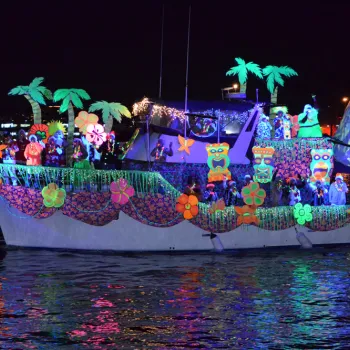 A boat decorated with neon tropical and floral designs glows at night, with costumed people on board, reflecting colorfully on the water.