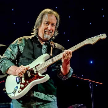 A man in a patterned shirt and vest plays an electric guitar on stage with a starry background and a partial sign visible behind him.