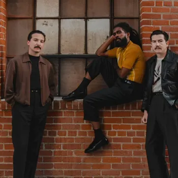 Three men pose against a red brick wall; one sits on a window ledge while the other two stand on either side, all wearing dark trousers and jackets or shirts.