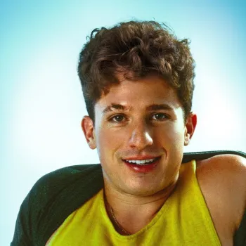 A young man with curly brown hair smiles at the camera, wearing a yellow tank top and a dark green jacket against a light blue background.