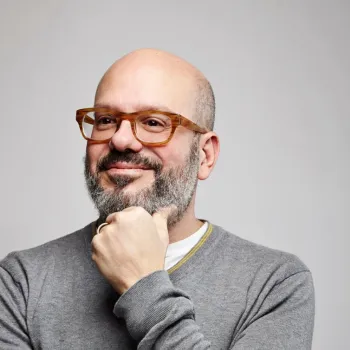 A bearded man with glasses and a gray sweater poses against a plain background, resting his chin on his hand and smiling slightly.