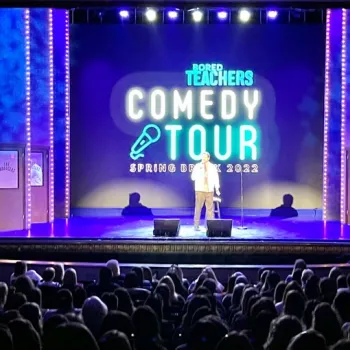 A comedian stands on stage performing at the Bored Teachers Comedy Tour, Spring Break 2022, in front of an audience in a theater.