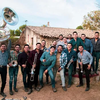 A group of musicians holding brass and percussion instruments pose outdoors in front of an old building with a tiled roof.