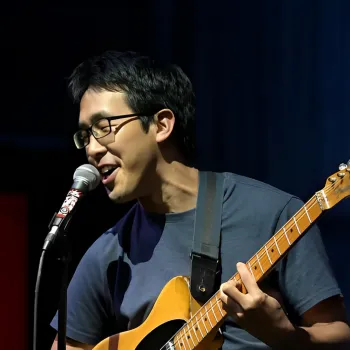 A person wearing glasses sings into a microphone while playing an electric guitar on stage.