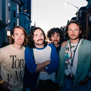 Four young men stand closely together on a residential street at dusk, facing the camera with neutral expressions.