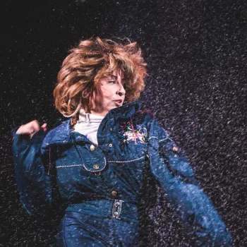 Person in a blue jumpsuit with embroidery dances energetically, hair flying, while water or confetti falls around them against a dark background.